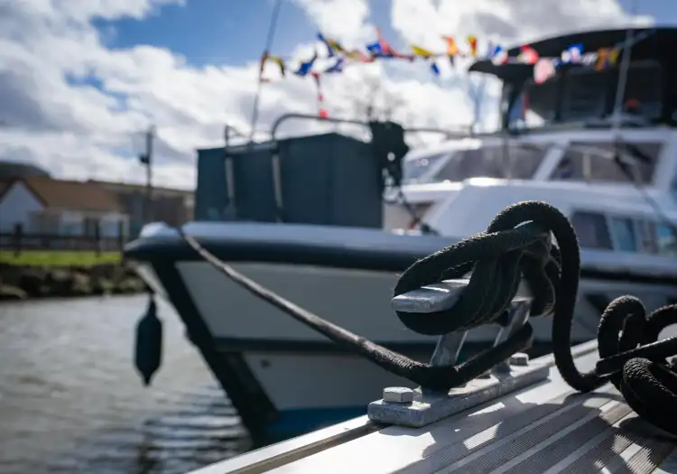 Gros plan d'un cordage noir noué à une bitte d'amarrage dans le port fluvial d'Aire-sur-la-Lys, avec des bateaux de plaisance amarrés en arrière-plan par beau temps