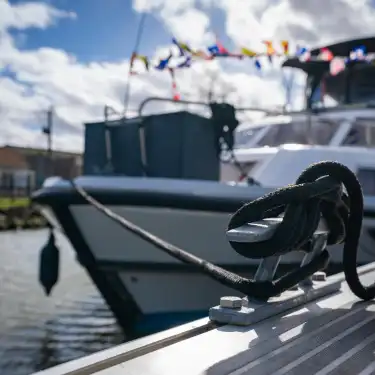 Gros plan d'un cordage noir noué à une bitte d'amarrage dans le port fluvial d'Aire-sur-la-Lys, avec des bateaux de plaisance amarrés en arrière-plan par beau temps