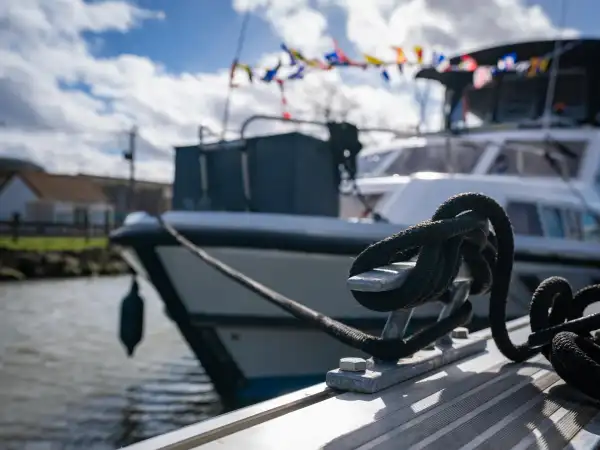 Gros plan d'un cordage noir noué à une bitte d'amarrage dans le port fluvial d'Aire-sur-la-Lys, avec des bateaux de plaisance amarrés en arrière-plan par beau temps